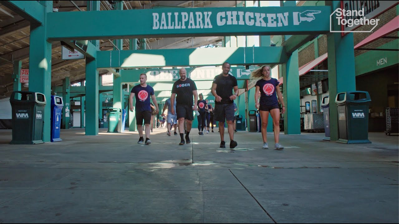 Group of people walking in a stadium concourse with signs, wearing shirts with various logos.