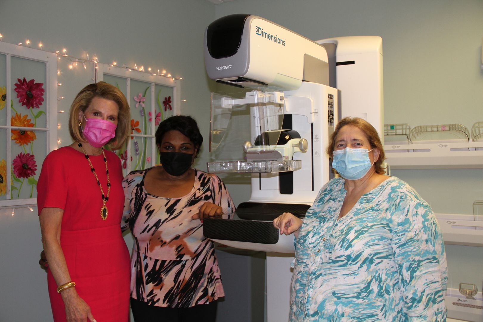 Three women wearing masks standing next to a "3Dimensions" medical machine in a room with floral decorations.