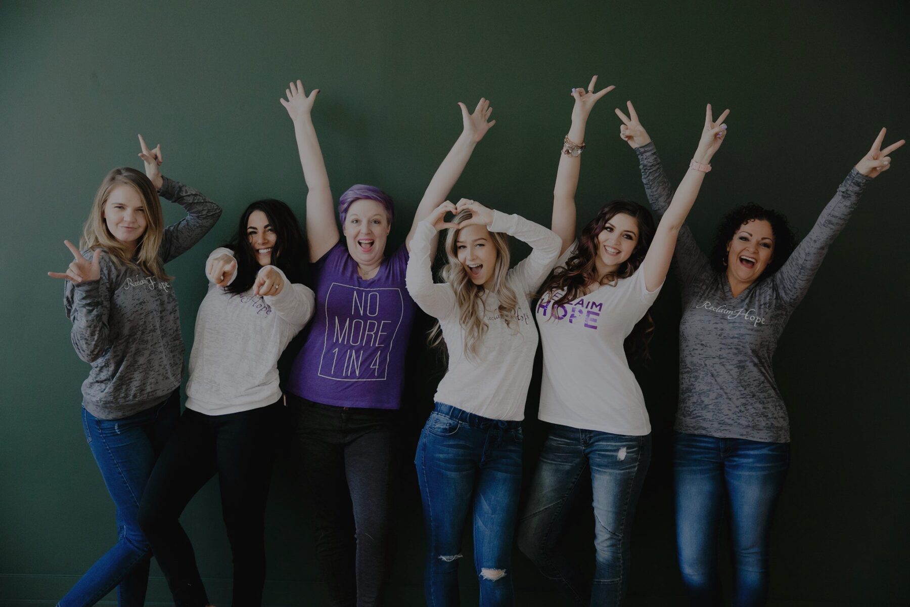 Group of six women posing joyfully in front of a green wall, some wearing shirts with positive messages.
