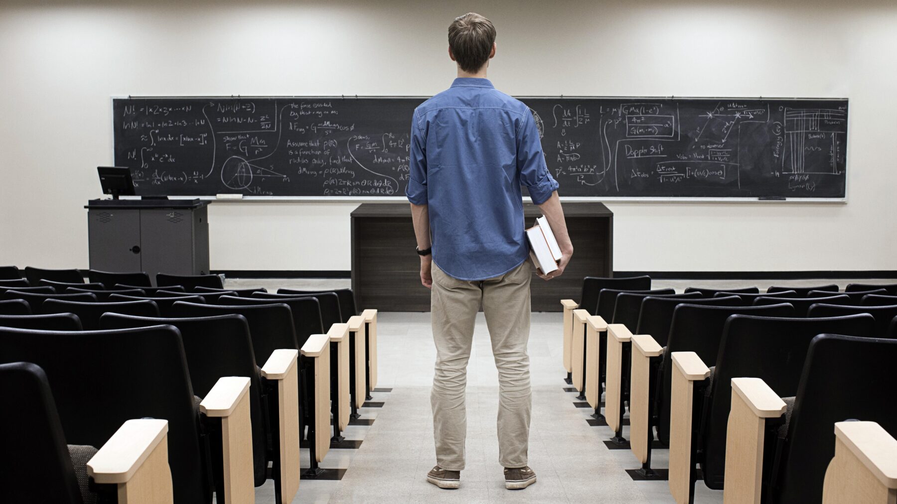 Man in a post-secondary classroom facing the board holding a book